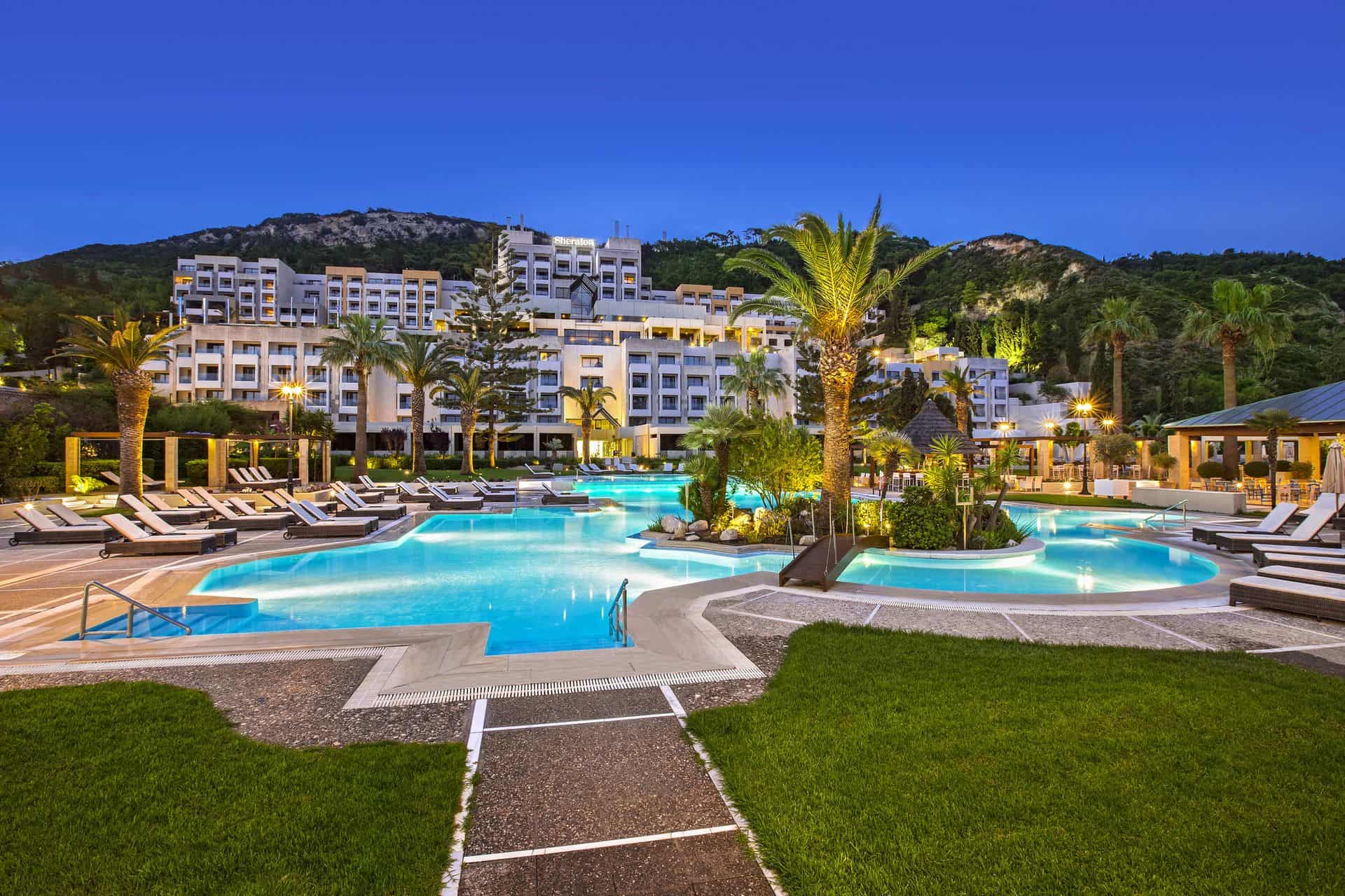 מלון Sheraton Rhodes בלילה - דקלים, דשא ירוק ומלון לבן על ההר